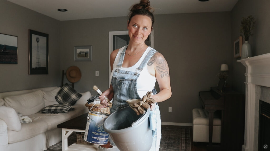 Christian homemaker wearing overalls and white tank top is holding a bucket and painting supplies